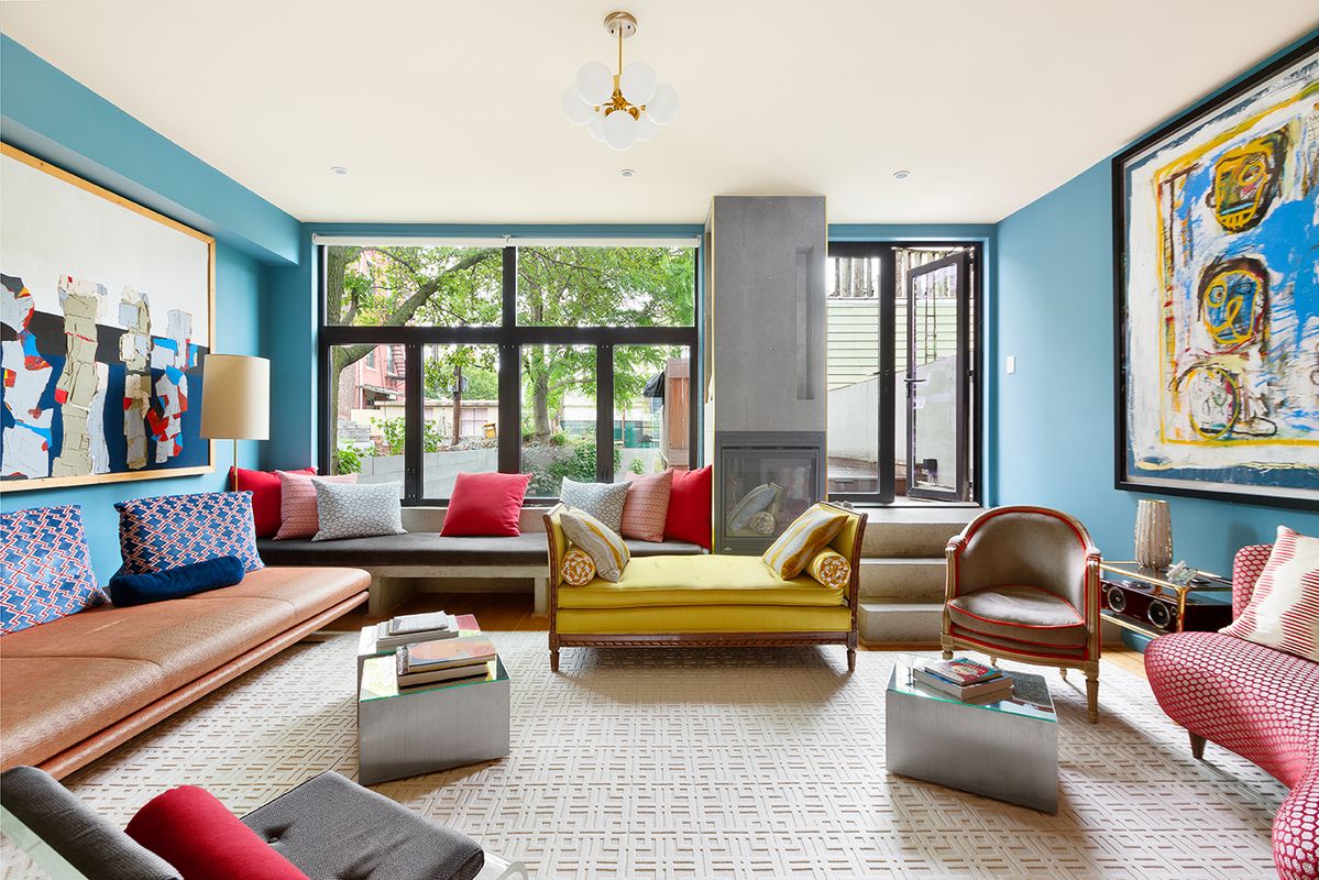 A living room with blue walls and unique furniture pieces - coffee table book on a cement side table, yellow love seat, large leather built in couch, and colorful paintings