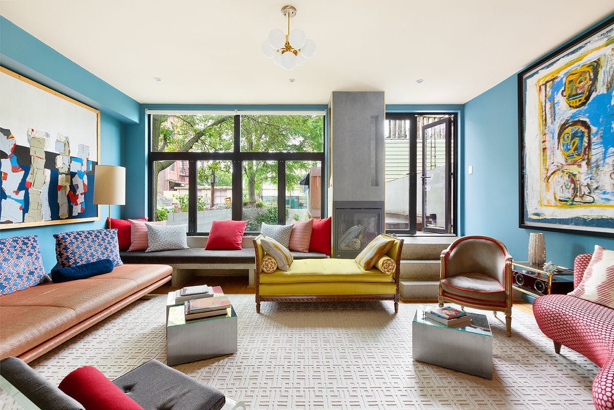A living room with blue walls and unique furniture pieces - coffee table book on a cement side table, yellow love seat, large leather built in couch, and colorful paintings 
